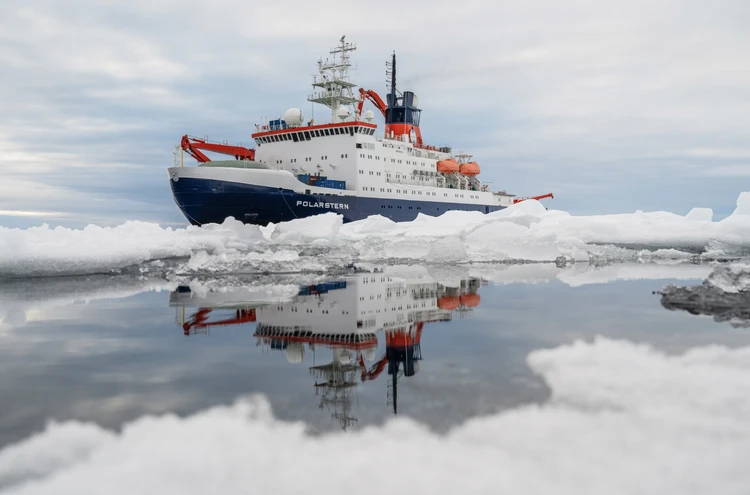 Zu sehen ist ein Eisbrecher, der durch eine eisige Polarlanschaft f&auml;hrt und sich im ruhigen Wasser spiegelt.
